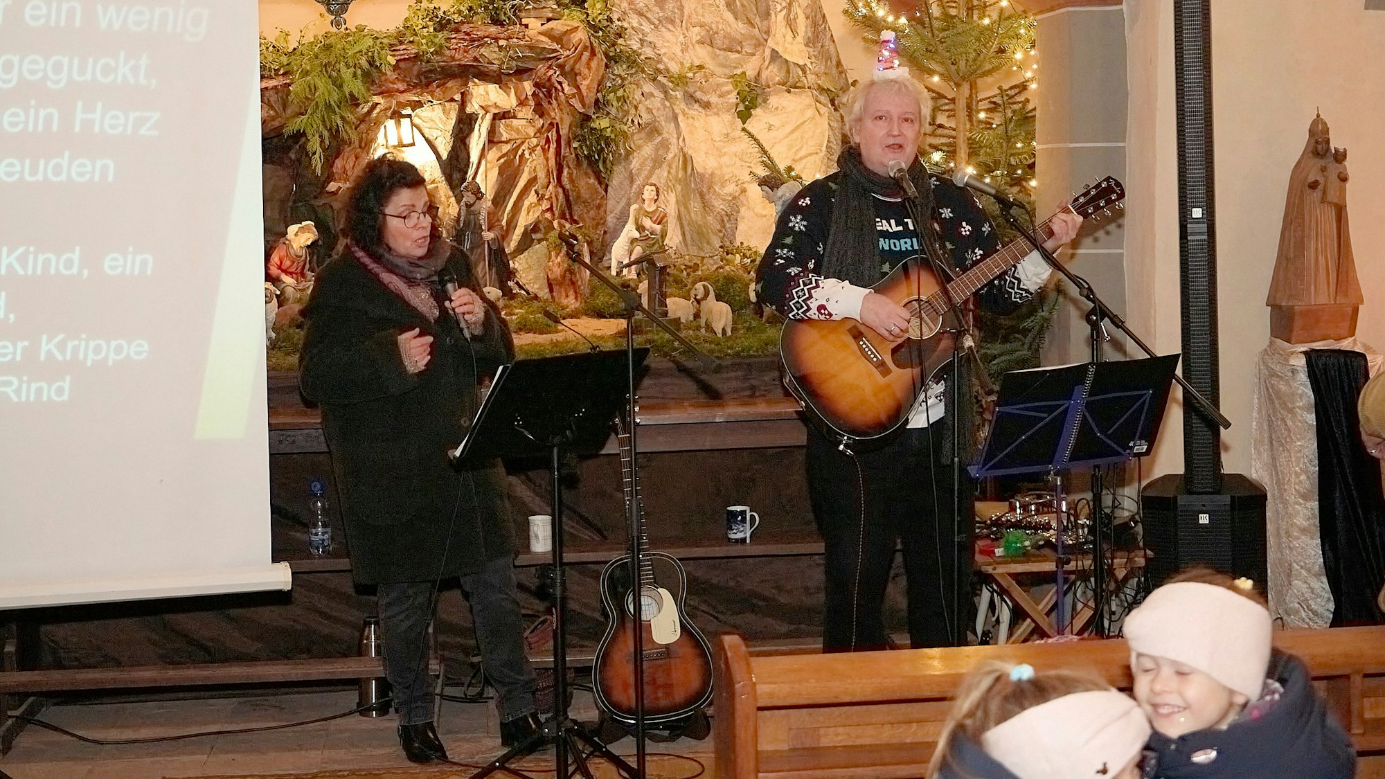 Eine Frau mit Mikrophon in der Hand und ein Mann, der Gitarre spielt, stehen im Altarraum einer Kirche. Hinter ihnen ist eine Weihnachtskrippe aufgebaut.