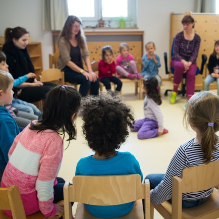 Kinder und Erzieherinnen sitzen in einer Kindertagesstätte in Düsseldorf (Nordrhein-Westfalen) im Kreis.