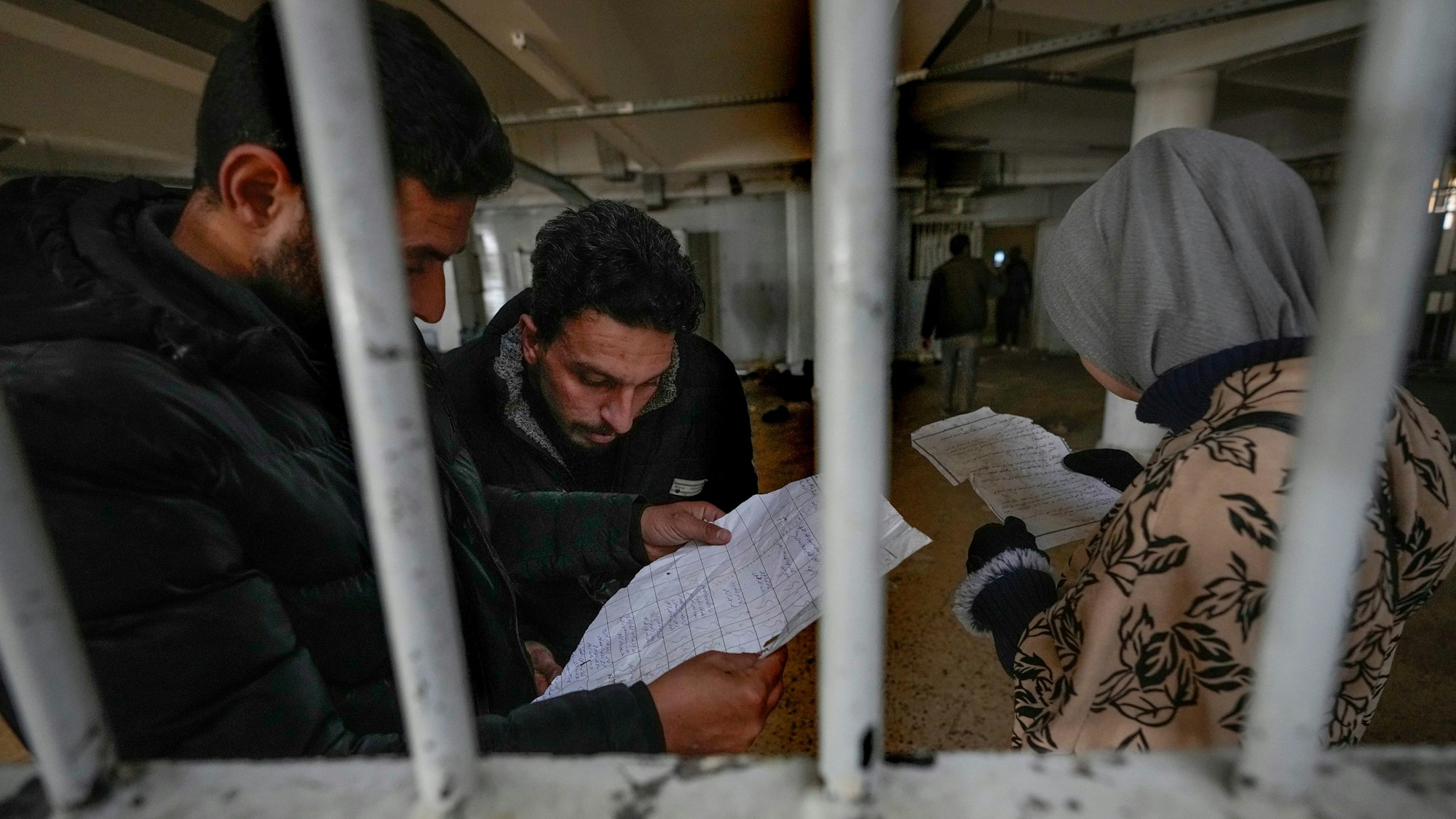 Zwei Männer und eine Frau, durch Gitterstäbe fotografiert, lesen zerknitterte Papiere.