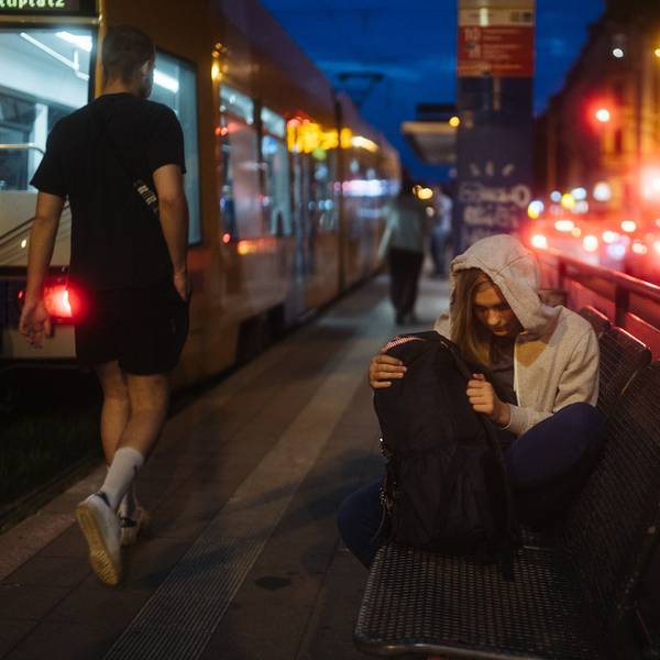 Eine Jugendliche mit Kapuzenpullover sitzt in der Nacht auf einer Bank in einer Großstadt. Neben ihr hält eine Straßenbahn.