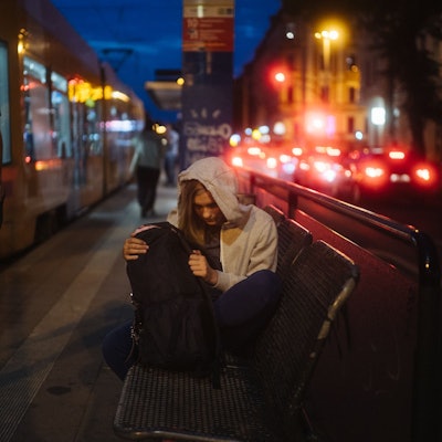 Eine Jugendliche mit Kapuzenpullover sitzt in der Nacht auf einer Bank in einer Großstadt. Neben ihr hält eine Straßenbahn.