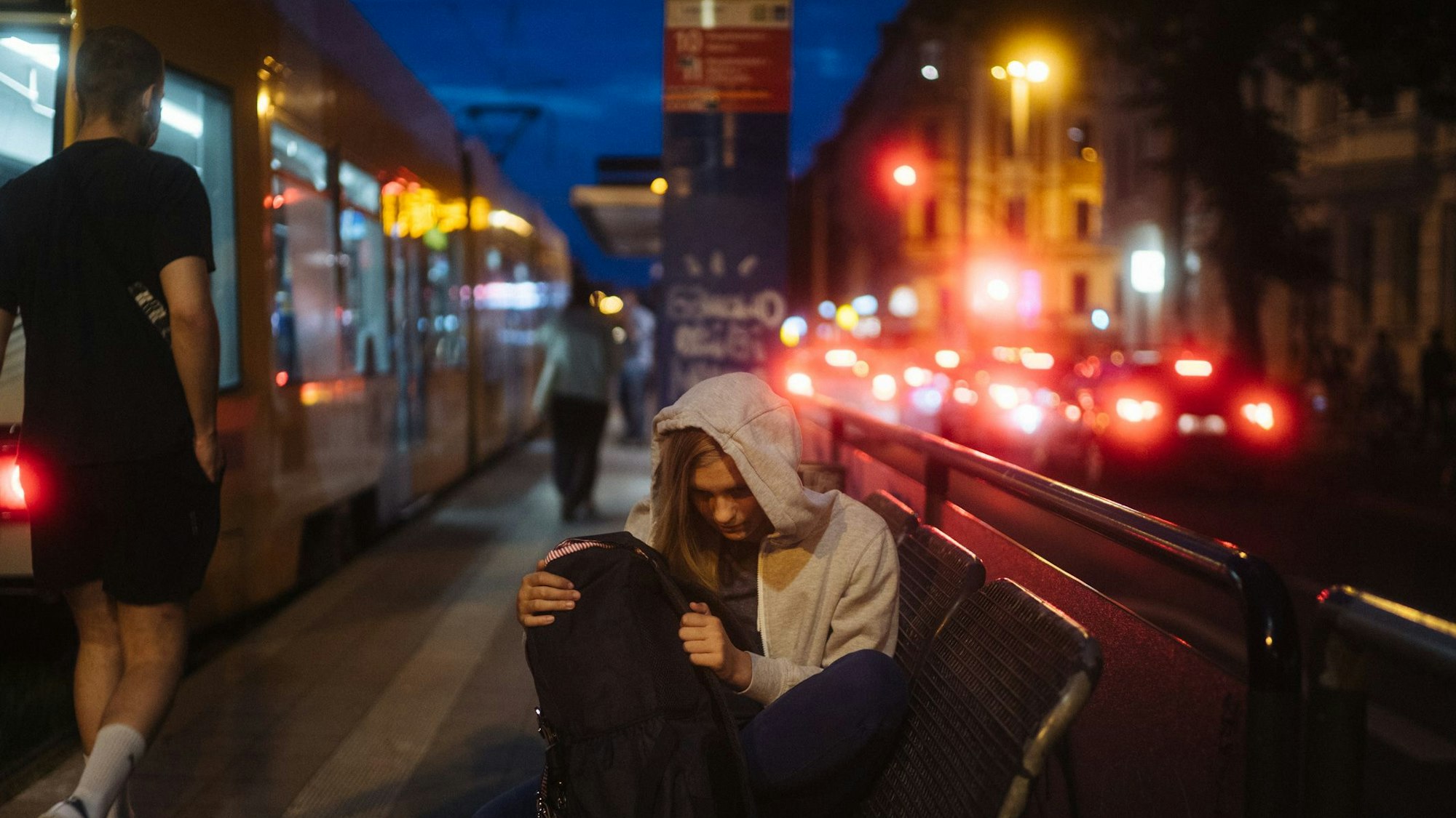 Eine Jugendliche mit Kapuzenpullover sitzt in der Nacht auf einer Bank in einer Großstadt. Neben ihr hält eine Straßenbahn.