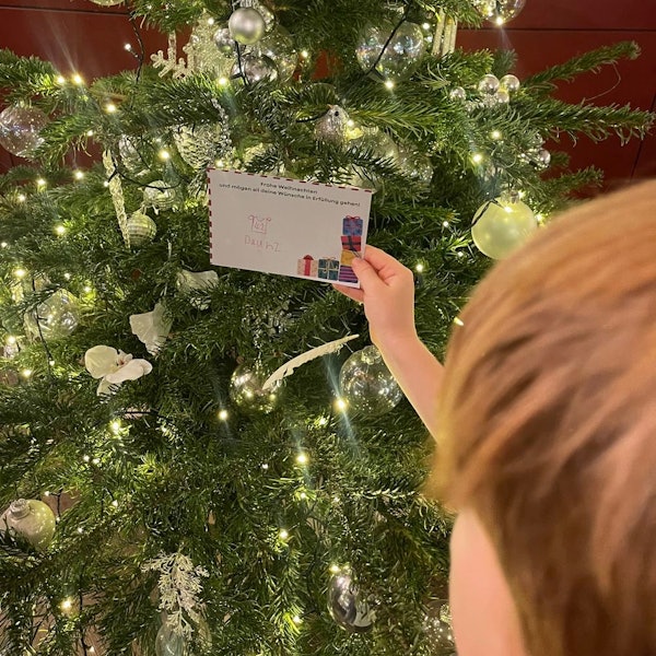 Ein Kind steht vor einem Tannenbaum und nimmt einen Wunschzettel ab, der daran gehängt ist.