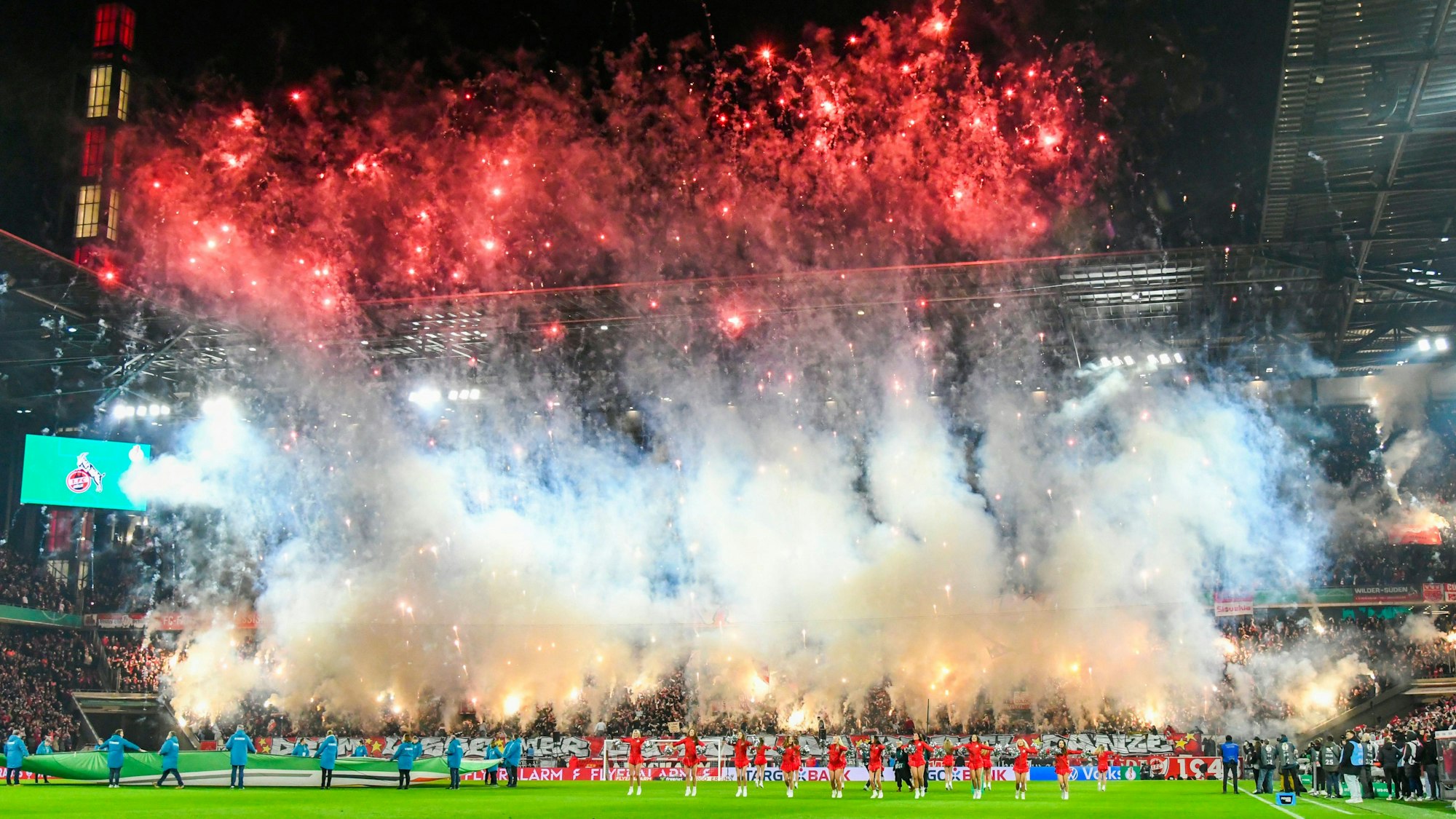 Während auf dem Rasen im Rhein-Energie-Stadion Köln Cheerleaderinnen vor der DFB-Pokal-Begegnung 1. FC Köln gegen Hertha BSC tanzen, brennen Fans große Mengen von Raketen und bengalischen Fackeln auf der Südtribüne ab.