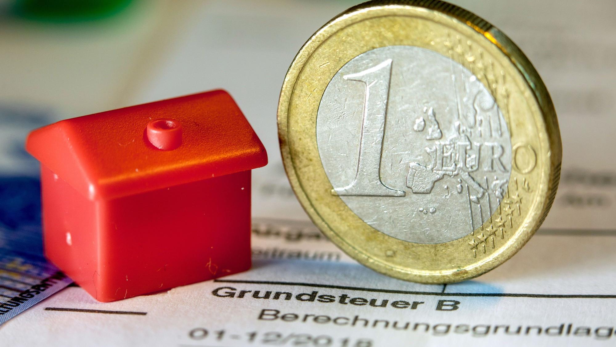 Eine Euro-Geldmünze sowie ein Spielzeughaus stehen auf einem Abgabenbescheid für die Entrichtung der Grundsteuer.