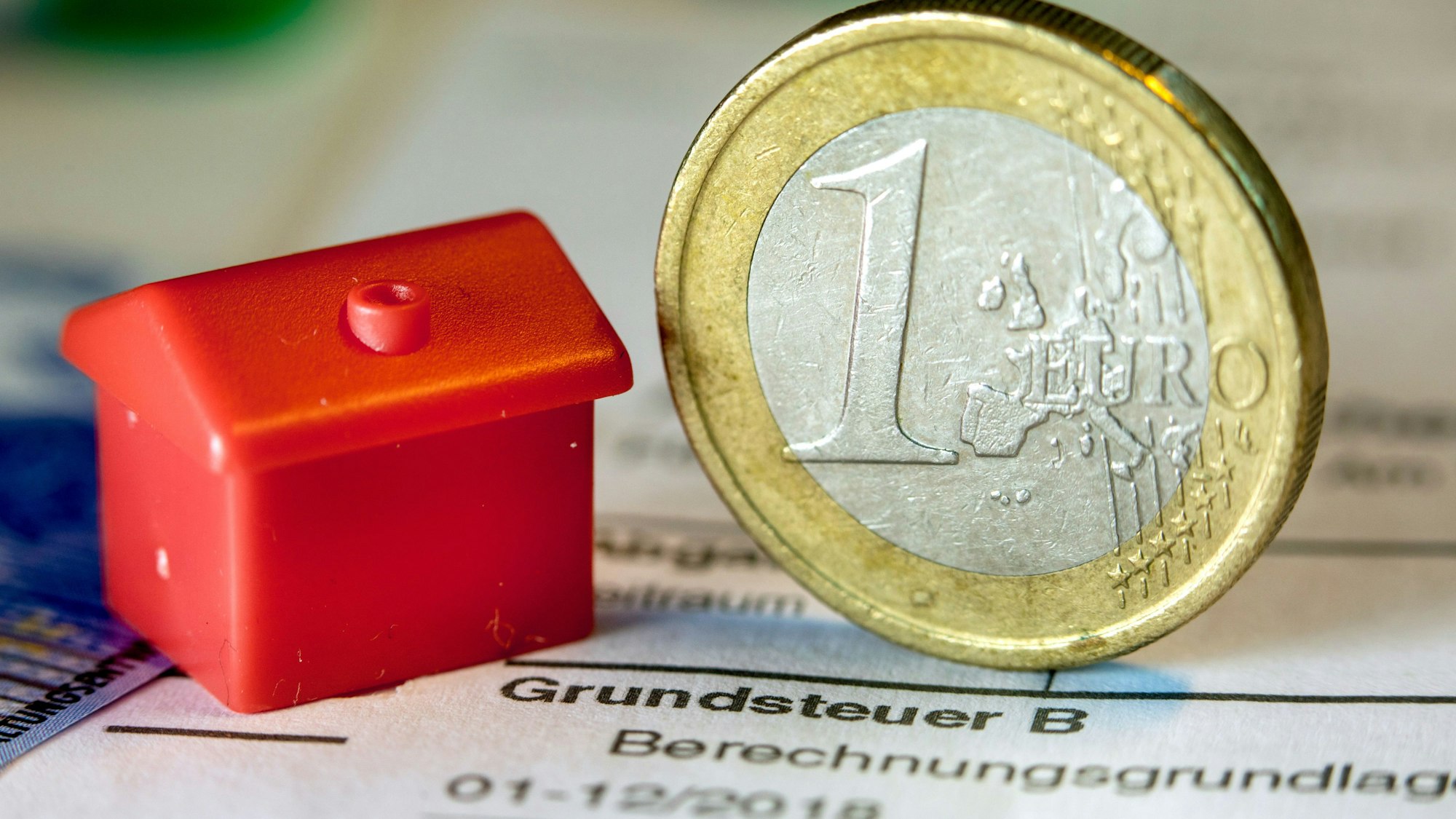 Eine Euro-Geldmünze sowie ein Spielzeughaus stehen auf einem Abgabenbescheid für die Entrichtung der Grundsteuer.