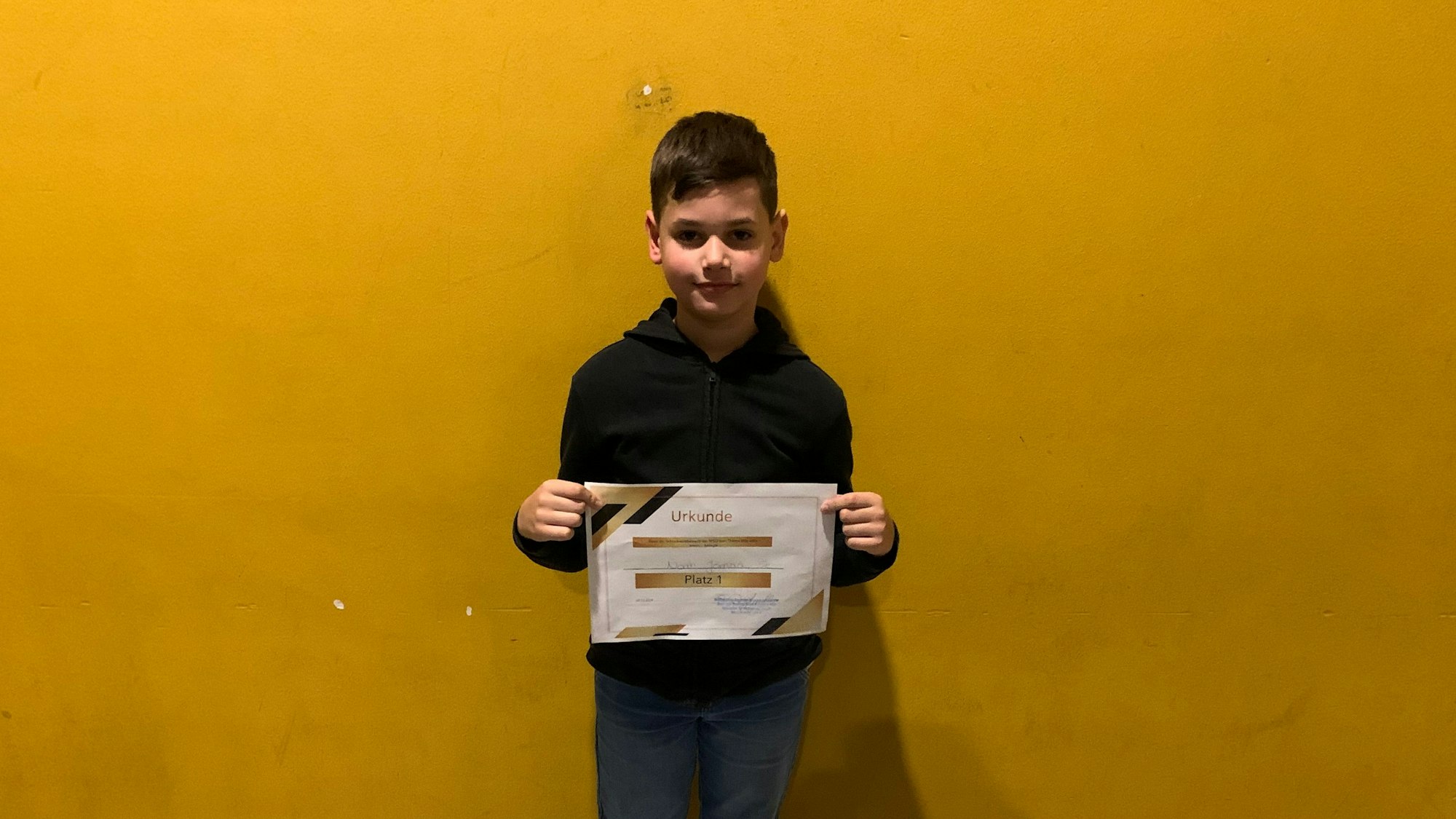 Ein Junge steht vor einer gelben Wand und zeigt eine Siegerurkunde.