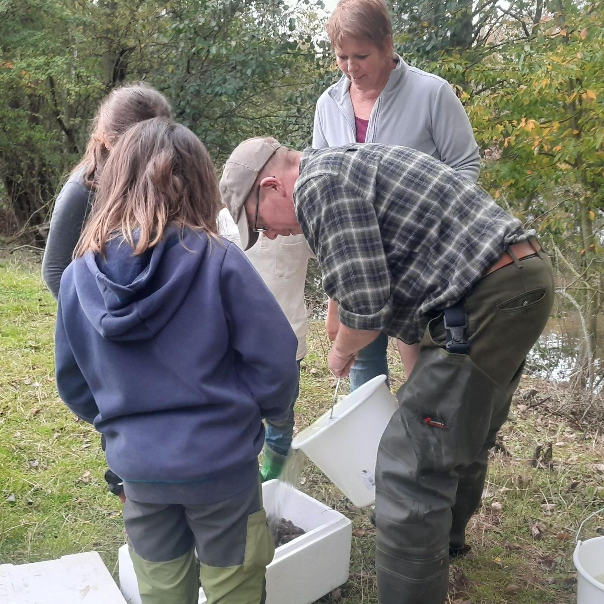 Ein Mann schüttet aus einem Eimer Wasser in einen Behälter, in dem sich Krebse befinden. Kinder und eine Frau schauen ihm dabei zu.