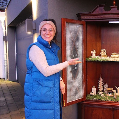 Christina Zinner steht neben dem Krippenschrank und zeigt auf die zwei Regale, auf denen hölzerne Figuren umgeben von Moos