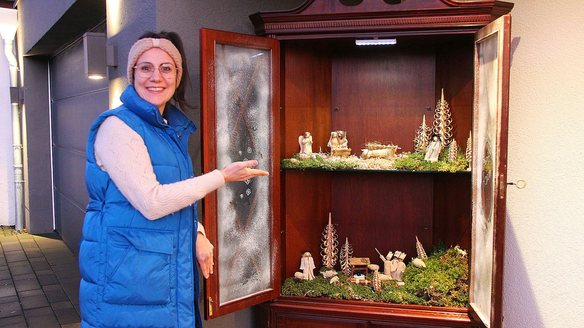 Christina Zinner steht neben dem Krippenschrank und zeigt auf die zwei Regale, auf denen hölzerne Figuren umgeben von Moos