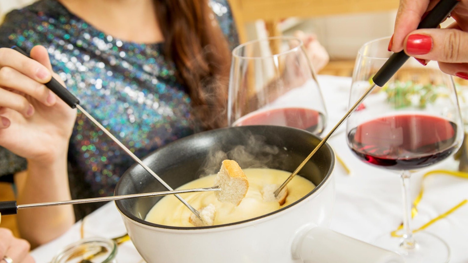 Ein Topf mit Käsefondue auf einem Tisch, dazu Rotwein und Brot an Spießen.