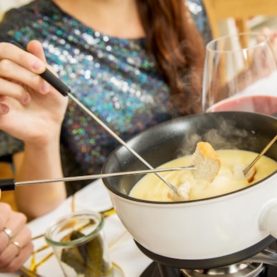 Ein Topf mit Käsefondue auf einem Tisch, dazu Rotwein und Brot an Spießen.