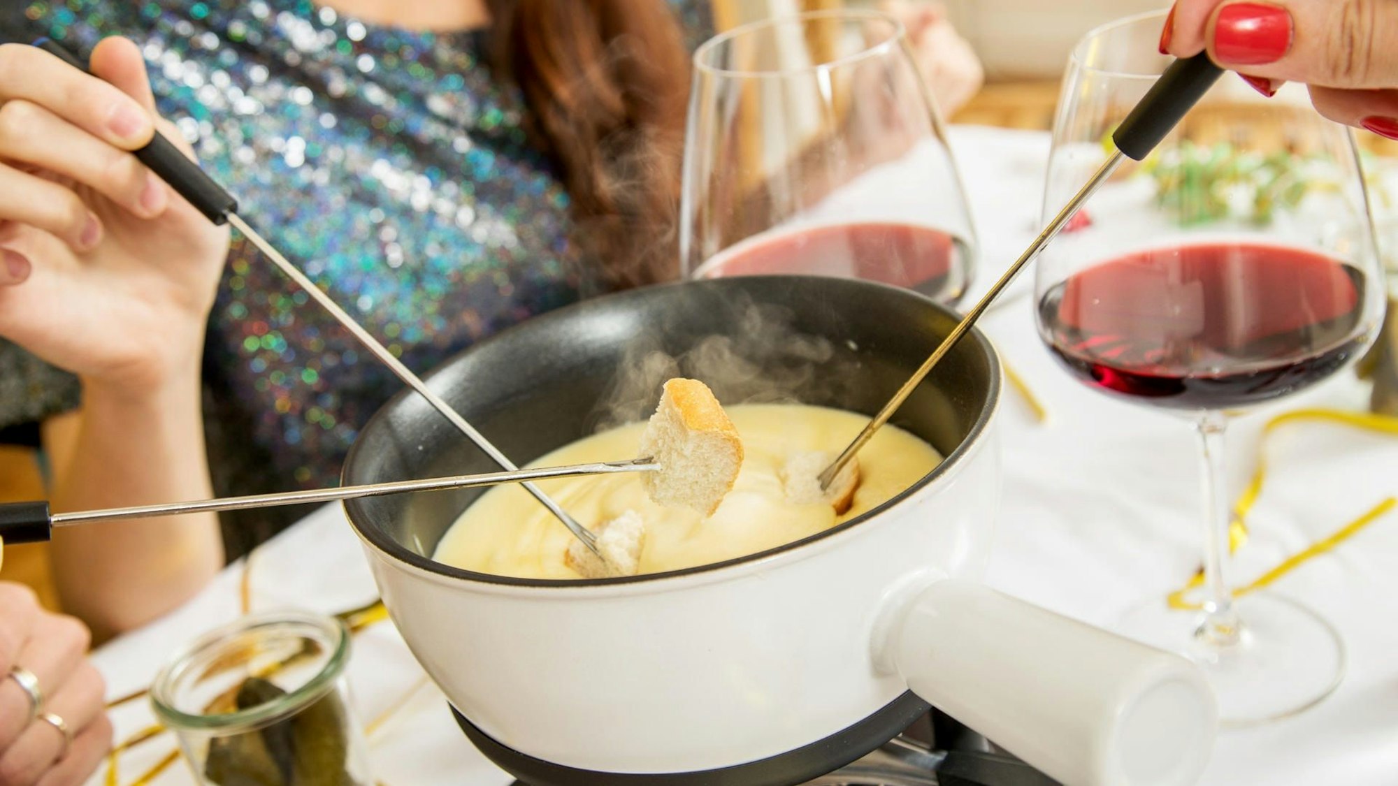 Ein Topf mit Käsefondue auf einem Tisch, dazu Rotwein und Brot an Spießen.