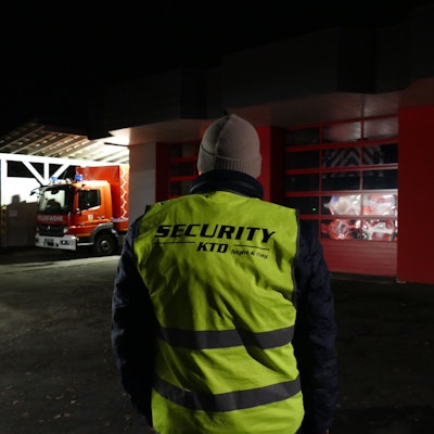 Ein Mann mit einer neongelben Weste, die mit dem Wort Security beschriftet ist, steht mit dem Rücken zur Kamera vor einem Feuerwehrgerätehaus.