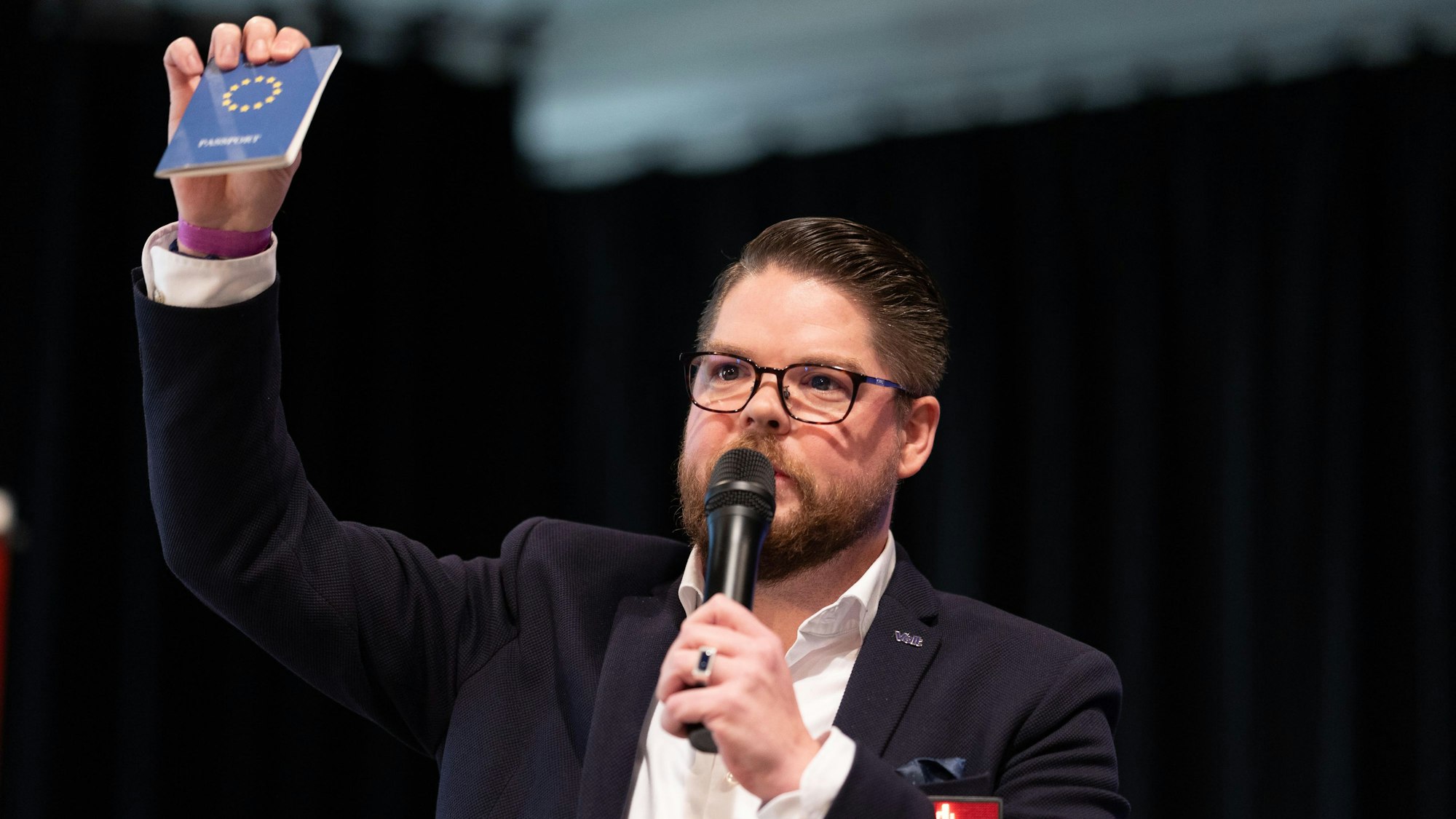 Ein Mann hält einen europäischen Pass und ein Mikrofon in der Hand.