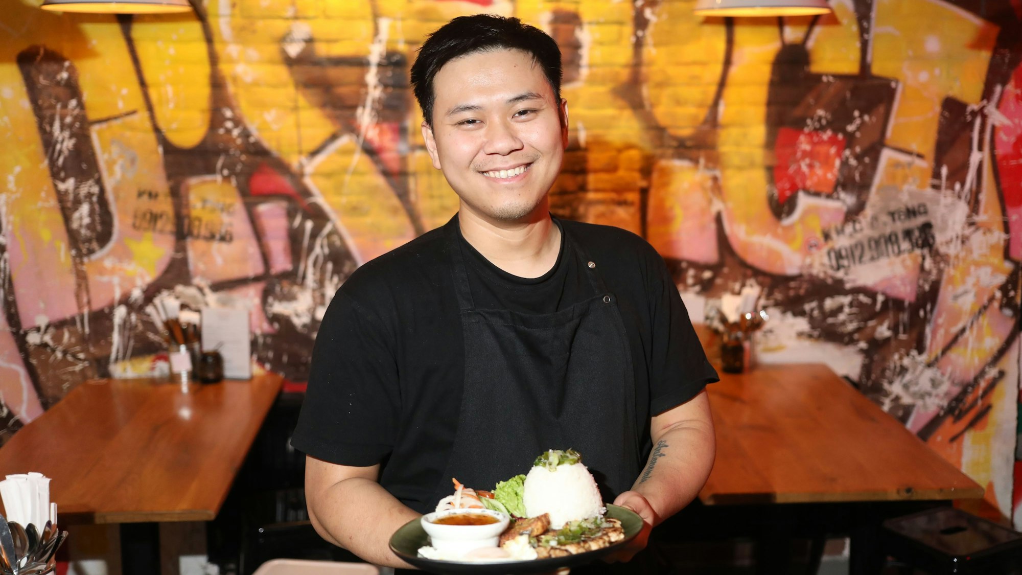 Inhaber Van Quan Nguyen ist mit einem Com Tam Tellergericht im Porträt zu sehen. Hinter ihm sieht man die Wand voller Graffiti.