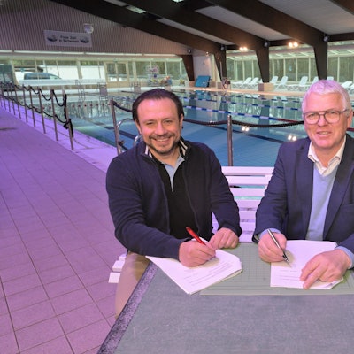 Zwei Männer sitzen in einem Hallenbad an einem Tisch und unterzeichnen einen Vertrag.