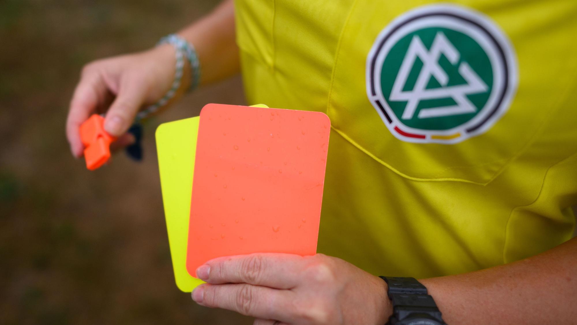 Ein Schiedsrichter steht mit roter und gelber Karte auf einem Rasen und hält in der anderen Hand eine Trillerpfeife.