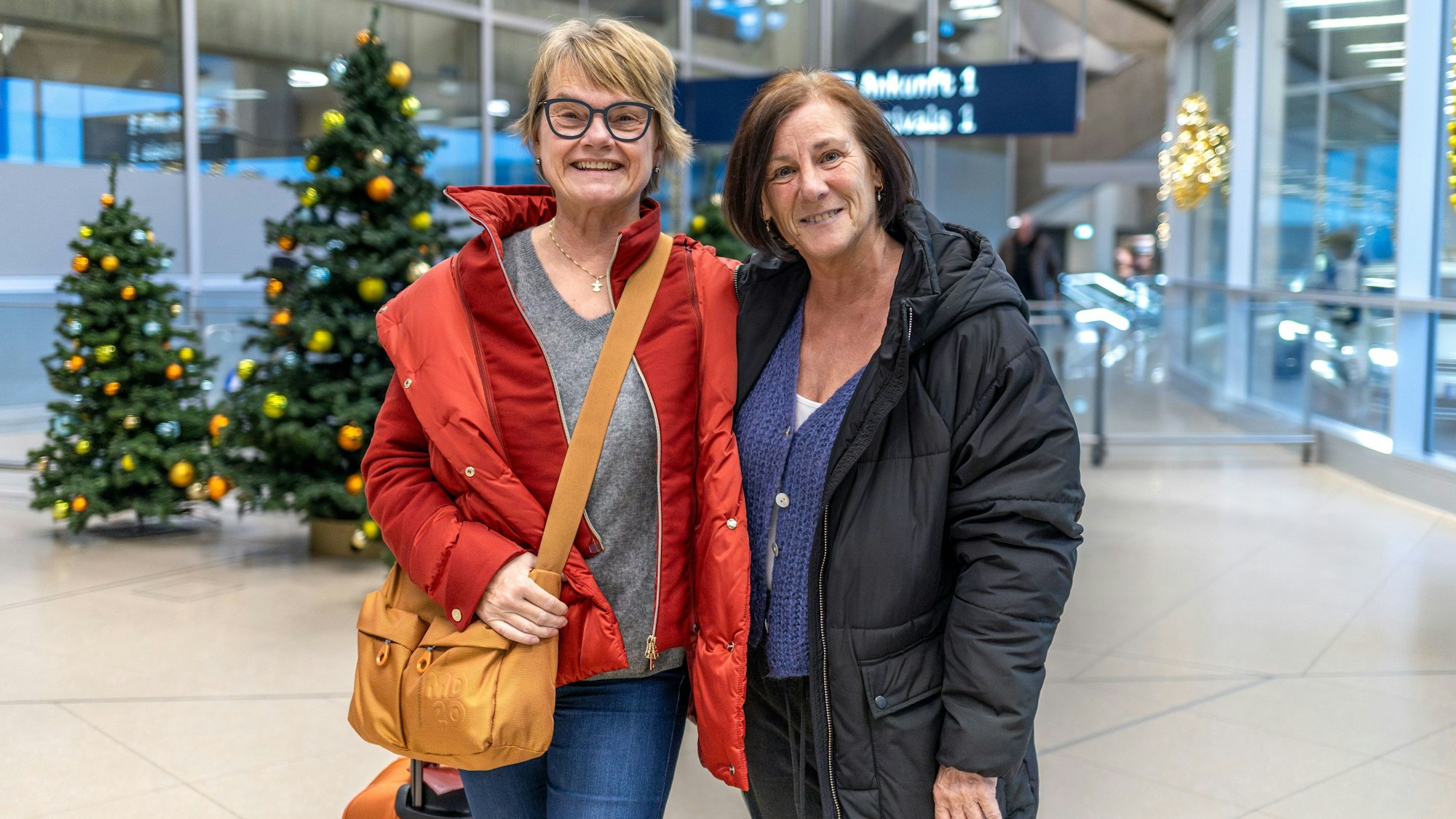 Zwei Frauen stehen Arm in Arm in der Ankunftshalle des Flughafens.