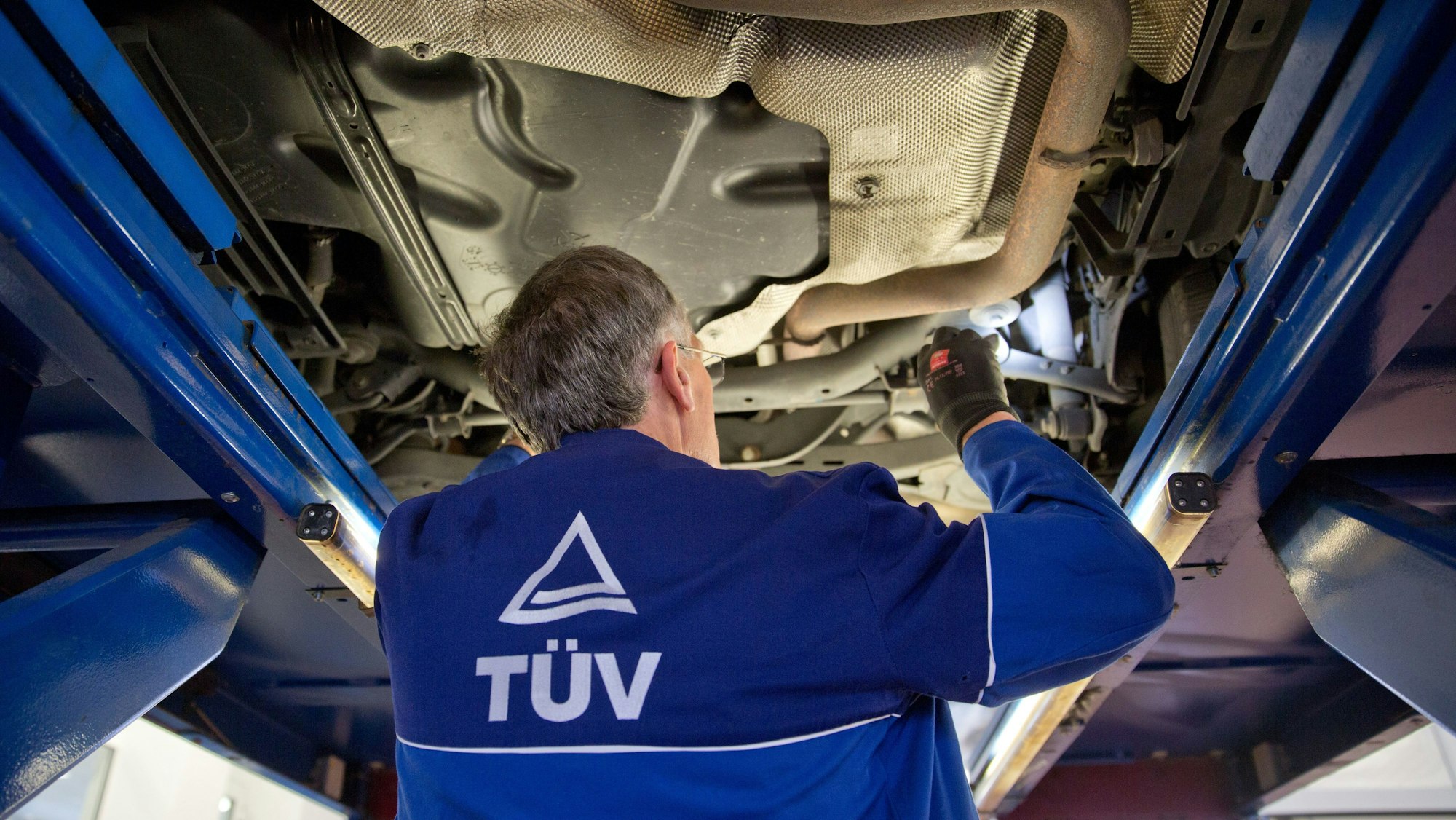 Ein Kfz-Sachverständiger vom Tüv überprüft den Unterboden eines aufgebockten Fahrzeugs. Der Sachverständige dreht dem Betrachter den Rücken zu. Er trägt eine Jacke mit dem Logo des Tüvs.