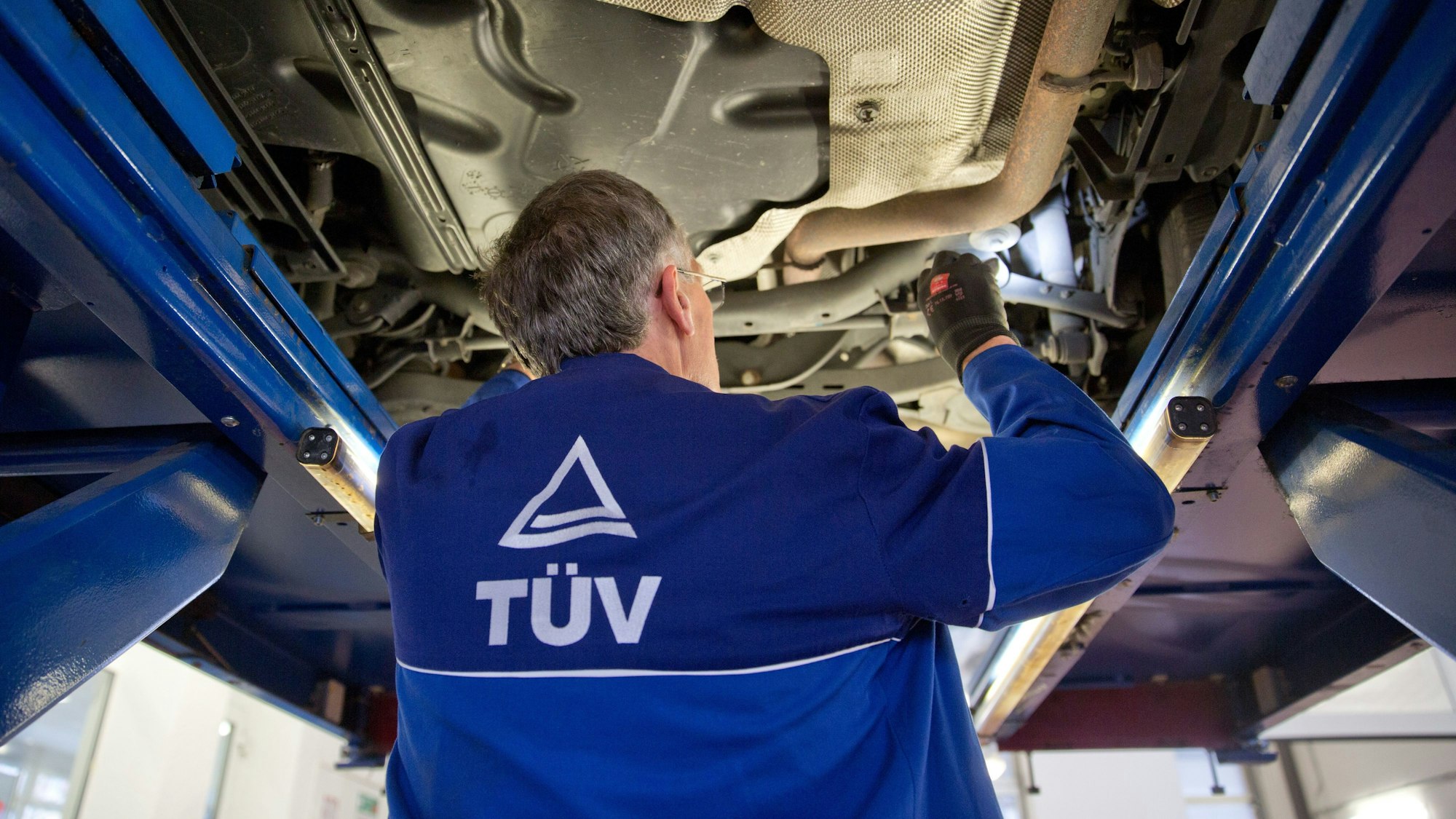 Ein Mann steht in einer Werkstatt unter einem Wagen und leuchtet die Unterseite mit einer Taschenlampe aus.