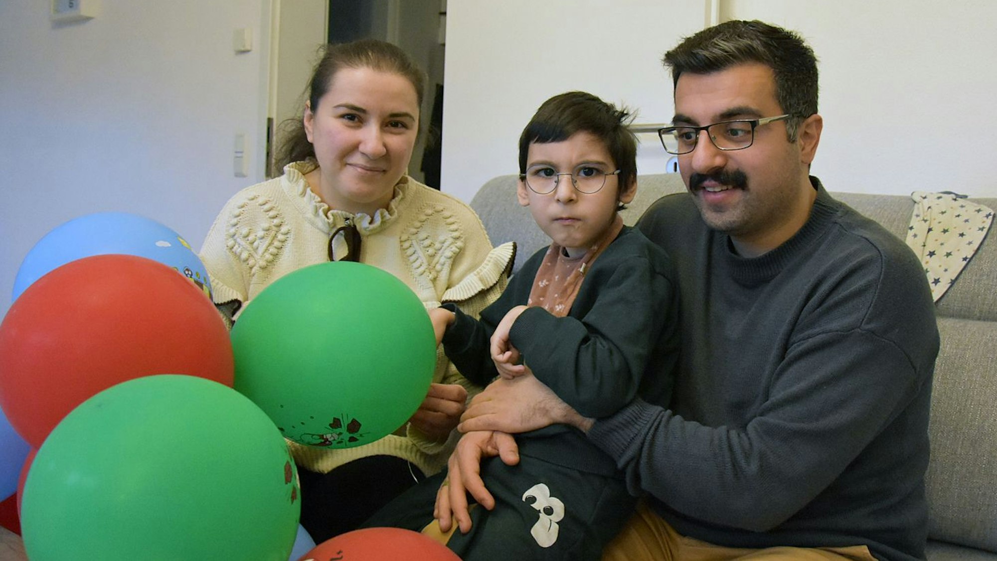 Ein Kind auf dem Schoß seines Vaters, daneben auf dem Sofa die Mutter, im Vordergrund bunte Ballons.