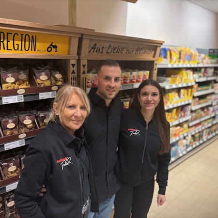 Auf dem Bild ist ein Mann mit seiner Mutter und seiner Freundin vor einem Supermarktregal zu sehen.
