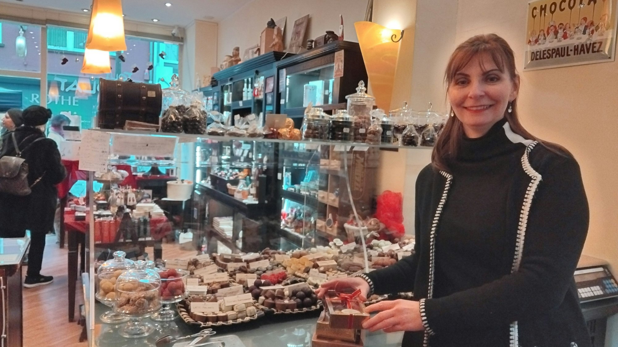 Eine Frau steht neben einer Pralinentheke in einem Schokoladengeschäft und hält ein als Geschenk verpacktes Pralinensortiment.