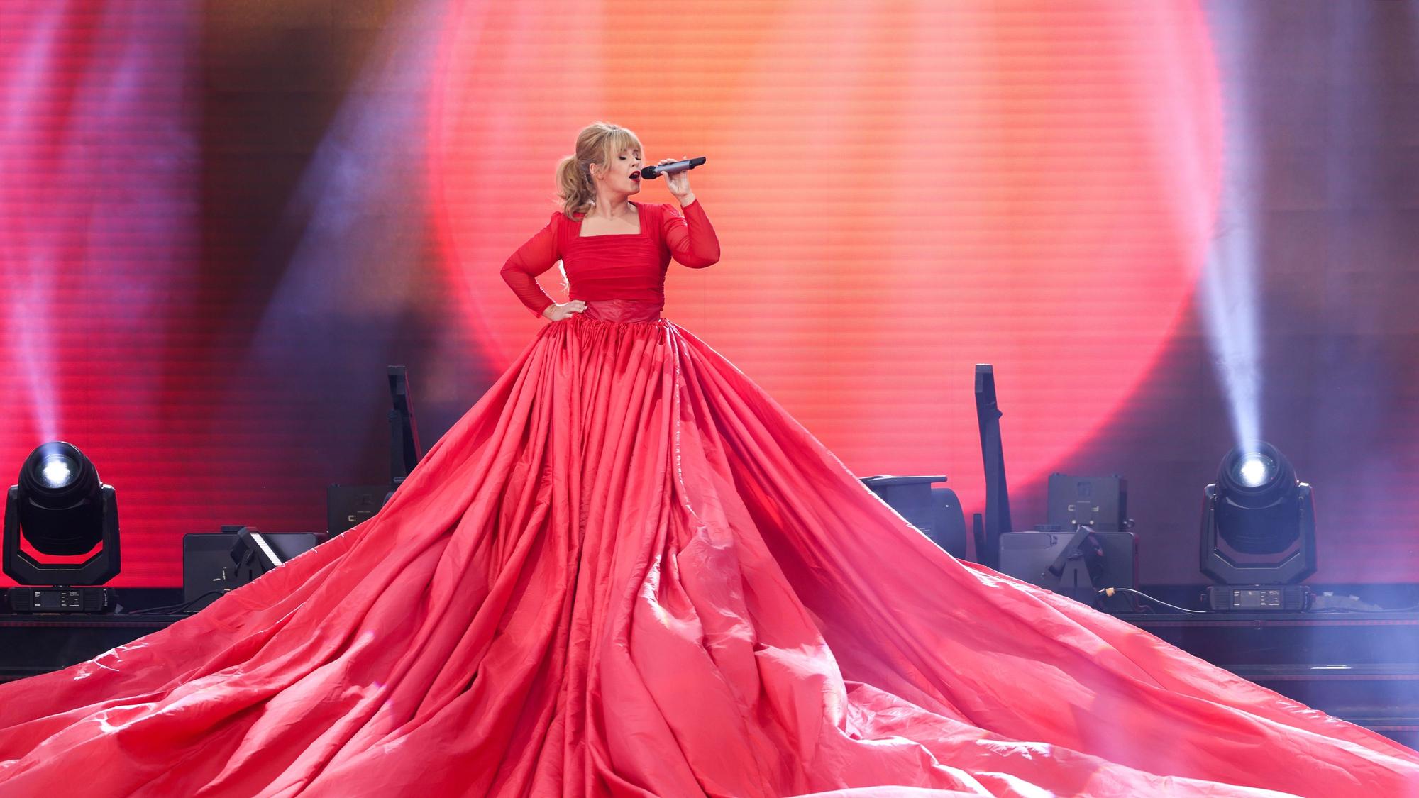 Maite Kelly steht auf einer Bühne. Sie trägt ein sehr langes pinkes Kleid und singt in ein Mikrofon.