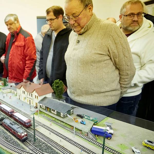 Um das Modell stehen mehrere Besucher. Sie blicken sehr interessiert auf die Züge, die Schienen und den Bahnhof im Kleinformat.