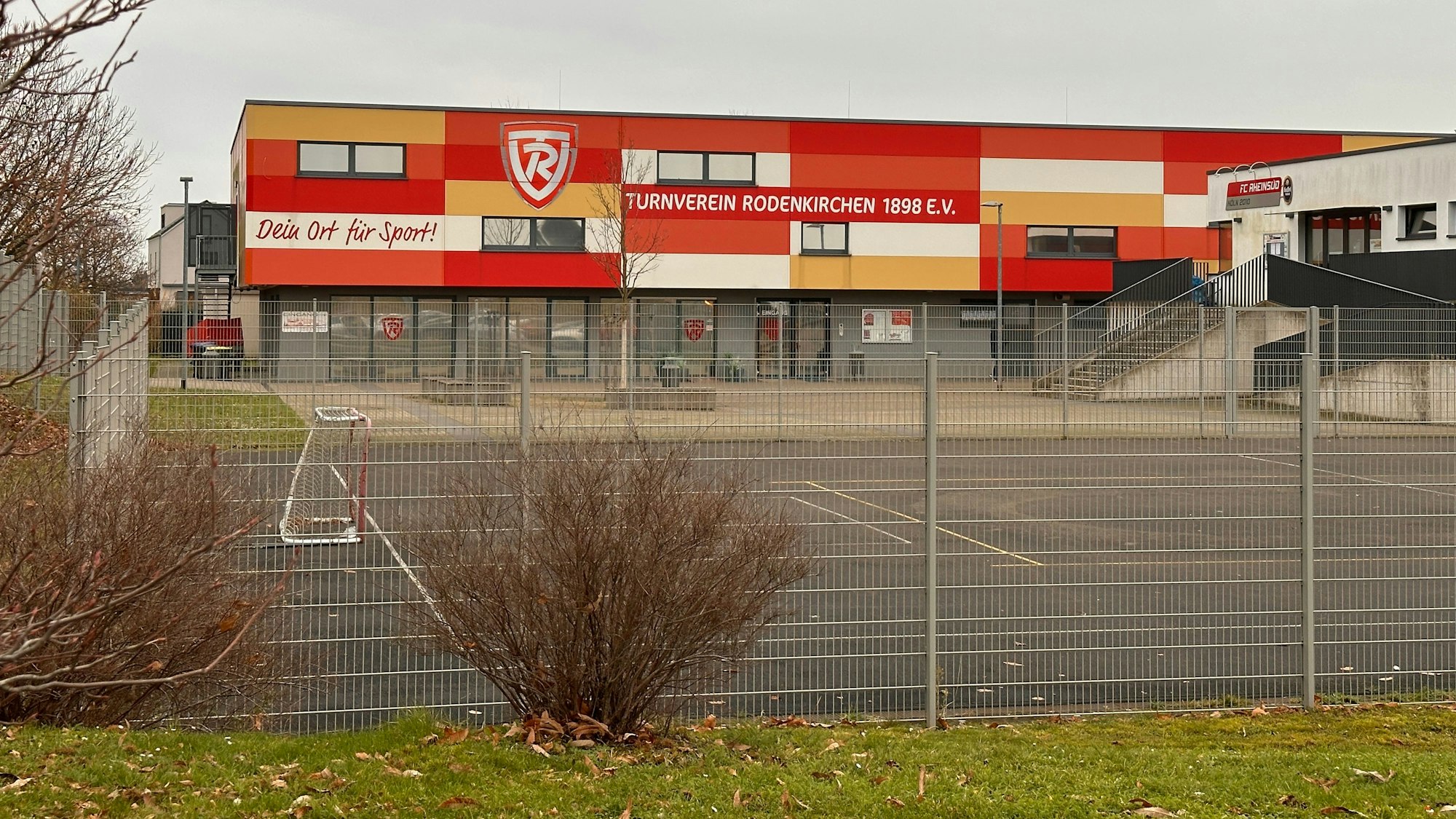 Zu sehen ist das umzäunte rot-orange-gelb-weiße Gebäude des Turnvereins Rodenkirchen.