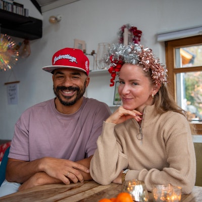 Marvin und Eva Becker sitzen an ihrem Esstisch. Marvin trägt eine rote Cappy mit der Aufschrift „Zuckerstange“ und Eva trägt einen Haarschmuck aus Lametta.