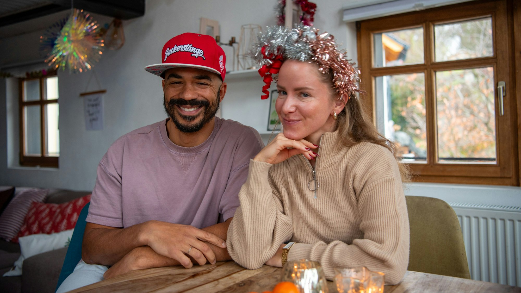 Marvin und Eva Becker sitzen an ihrem Esstisch. Marvin trägt eine rote Cappy mit der Aufschrift „Zuckerstange“ und Eva trägt einen Haarschmuck aus Lametta.