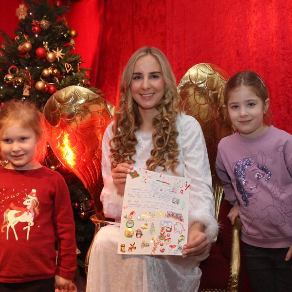 Zwei Kinder stehen links und rechts neben einer Christkinddarstellerin, die auf einem goldenen Stuhl sitzt und Engelsflügel trägt.