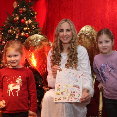 Zwei Kinder stehen links und rechts neben einer Christkinddarstellerin, die auf einem goldenen Stuhl sitzt und Engelsflügel trägt.