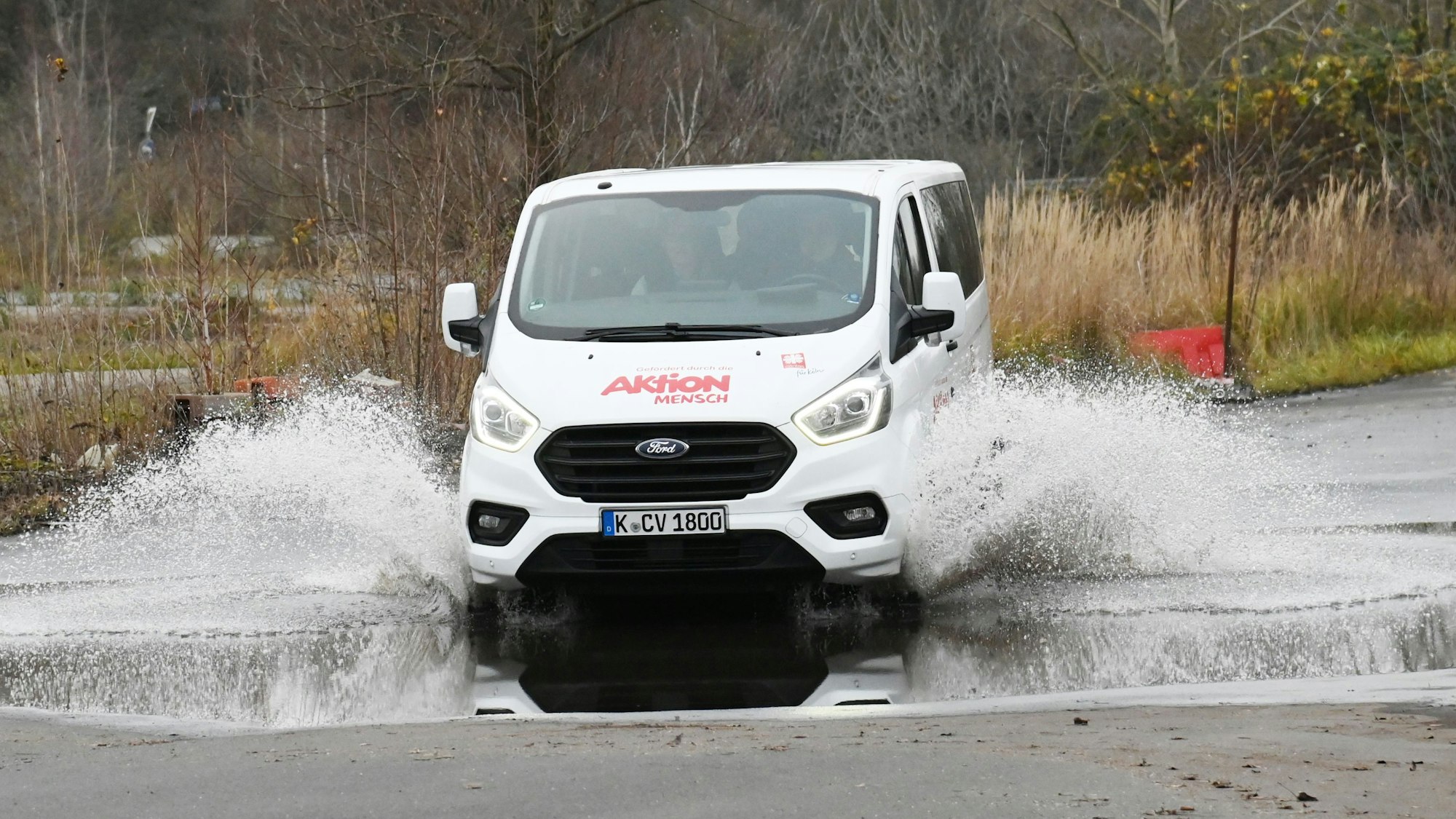 Ein weißer Bulli von „Aktion Mensch“ fährt durch eine große Pfütze.