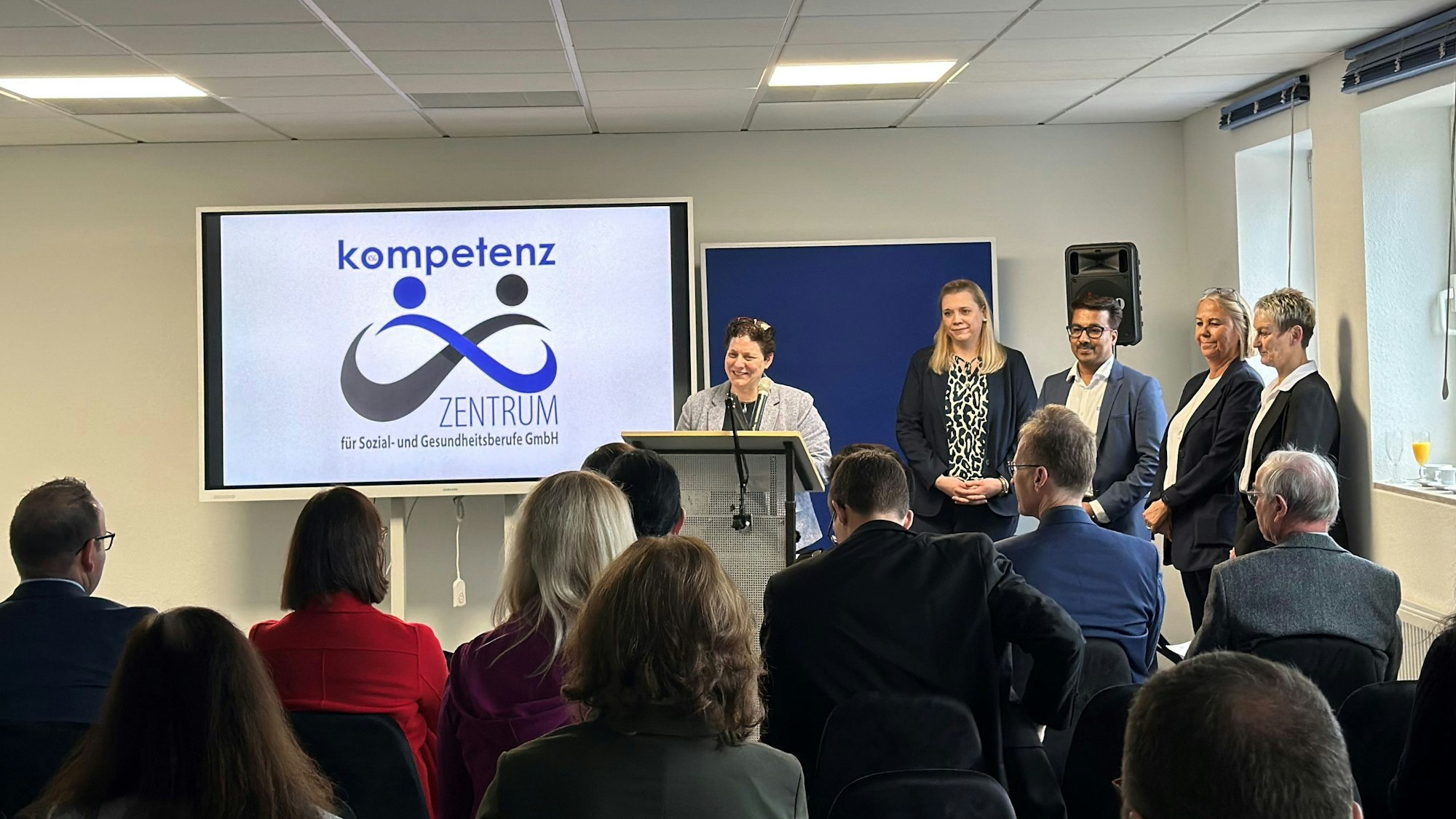 Eine Frau steht an einem Rednerpult, davor sitzen Zuhörer.