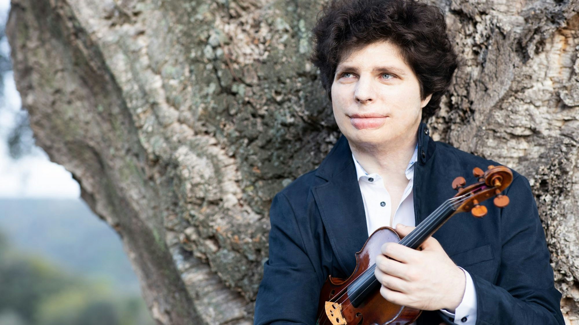 Augustin Hadelich hält, an einen Baum gelehnt, seine Geige