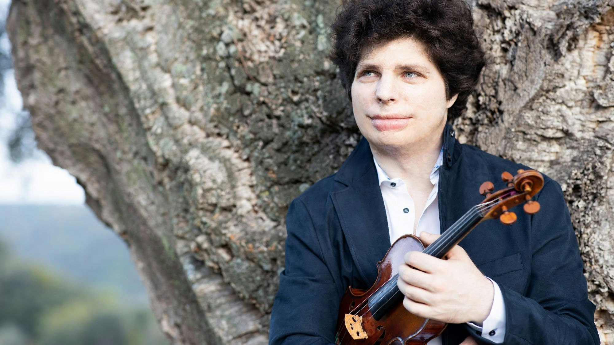 Der Geigen-Virtuose Augustin Hadelich steht vor einem Baum.
