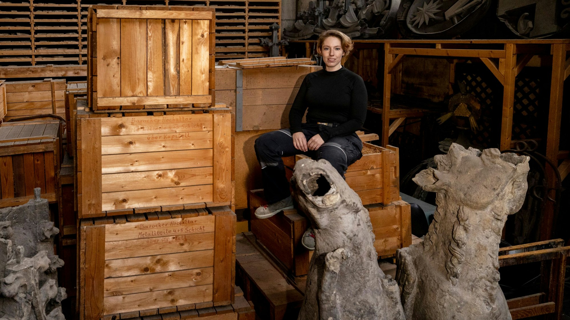 Steinmetzin Tanja Pinkale von der Kölner Dombauhütte sitzt in einem Depot auf dem sogenannten Uhrenboden des Doms. Vor ihr zwei stark verwitterte Wasserspeier vom mittelalterlichen Chor.