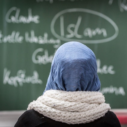 Eine Schülerin mit Kopftuch sitzt in einer Schule bei einer Unterrichtsstunde zum Thema Islam vor einer Tafel.