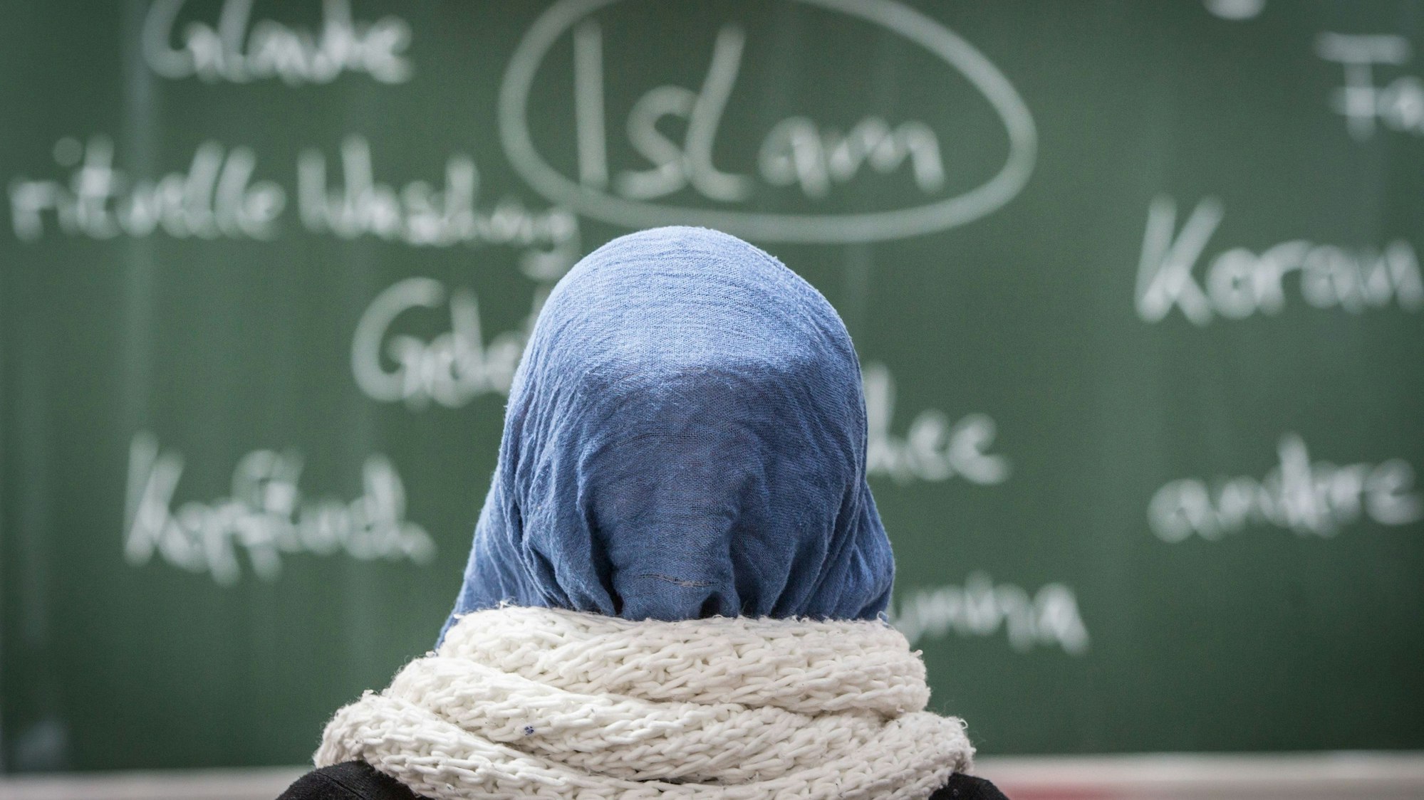 Eine Schülerin mit Kopftuch sitzt in einer Schule bei einer Unterrichtsstunde zum Thema Islam vor einer Tafel.