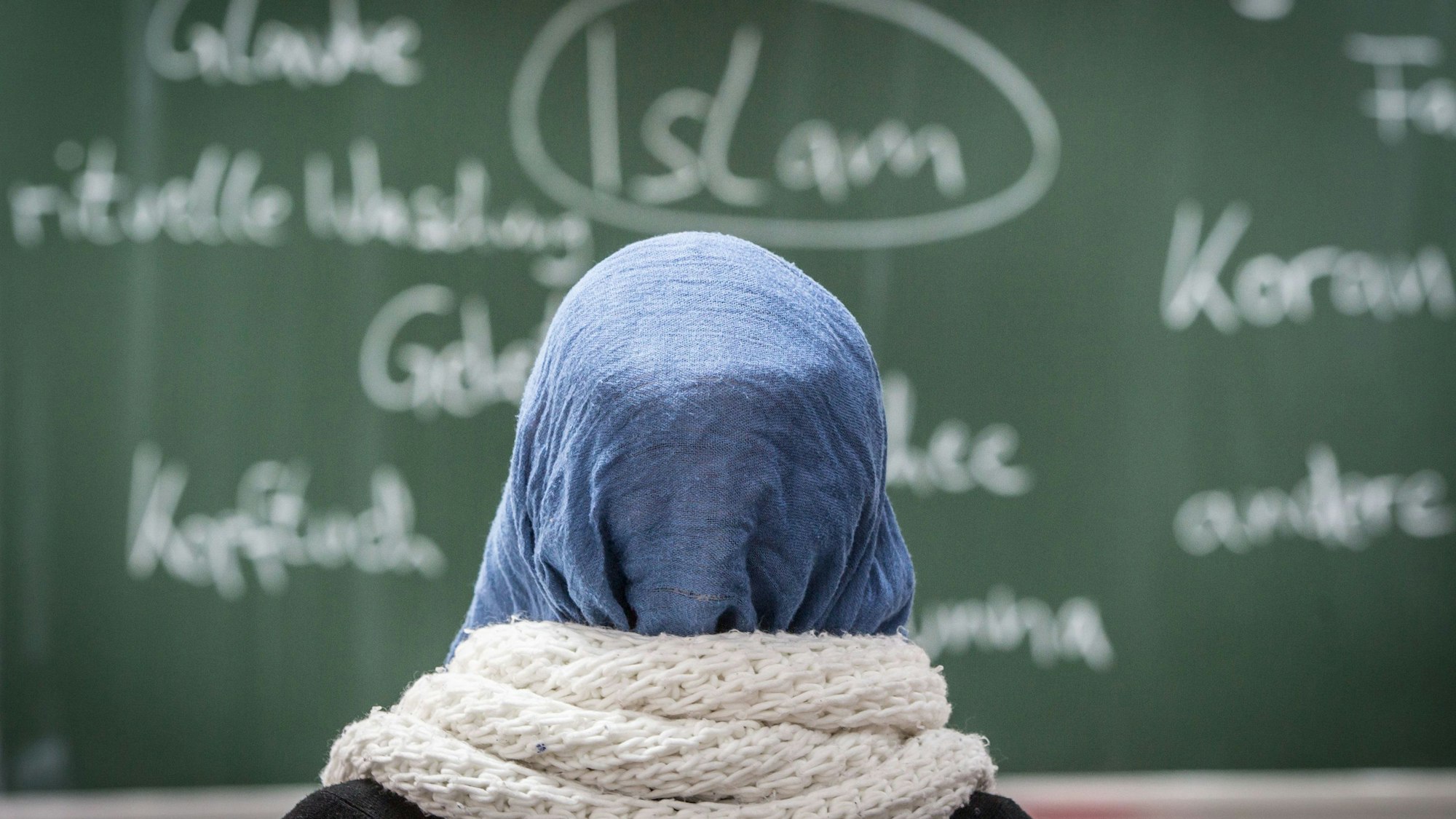 Eine Schülerin mit Kopftuch sitzt in einer Schule bei einer Unterrichtsstunde zum Thema Islam vor einer Tafel.