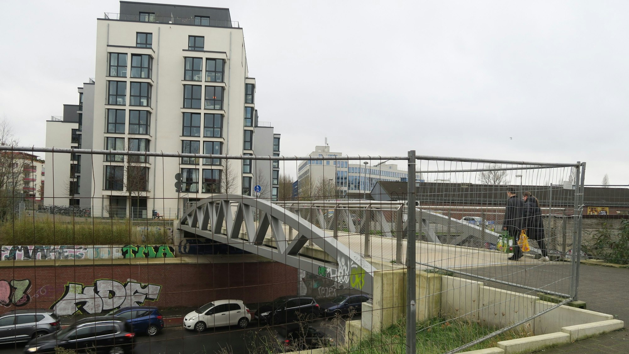 Blick auf eine Fußgängerbrücke, darunter fahren Autos, im Vordergrund steht ein Bauzaun.