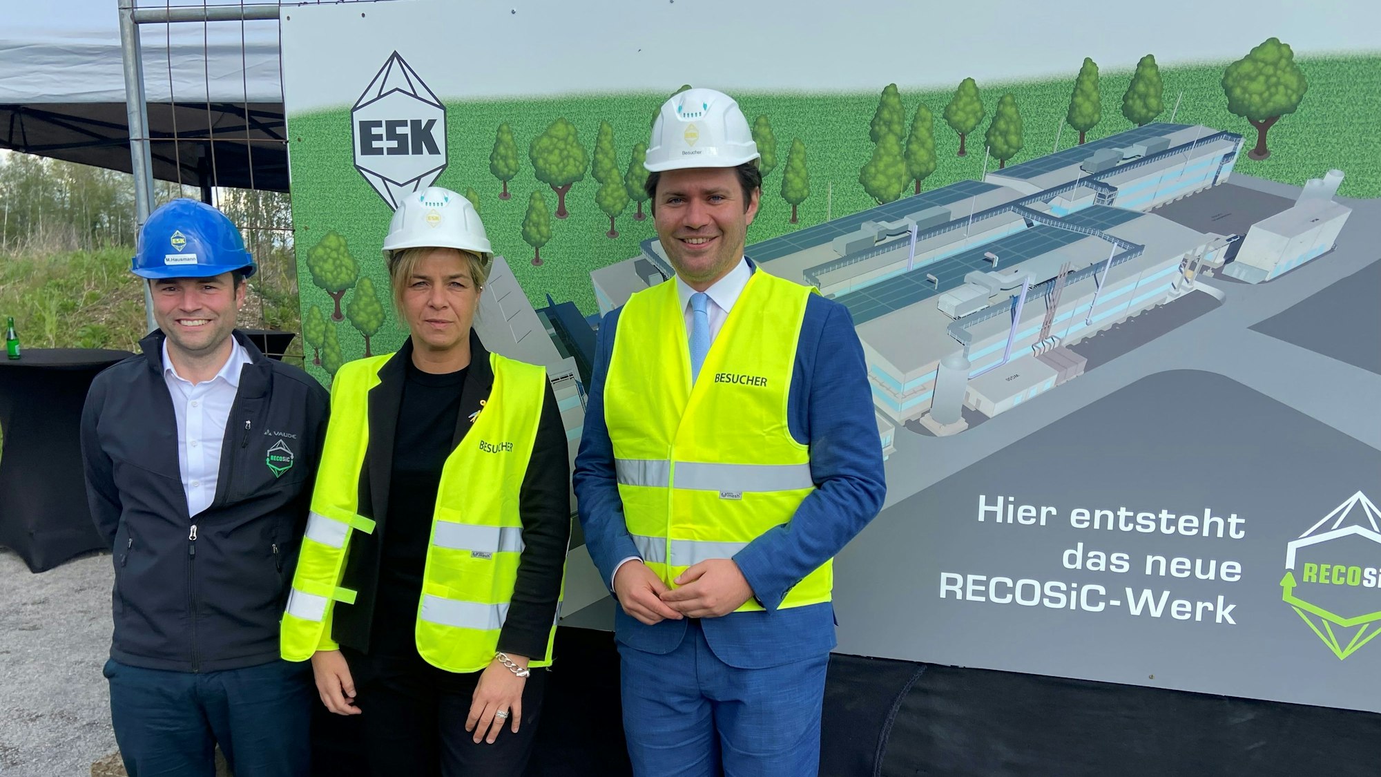 Auf dem Bild sind zwei Männer und eine Frau mit Sicherheitswesten vor einem Bauschild zu sehen.