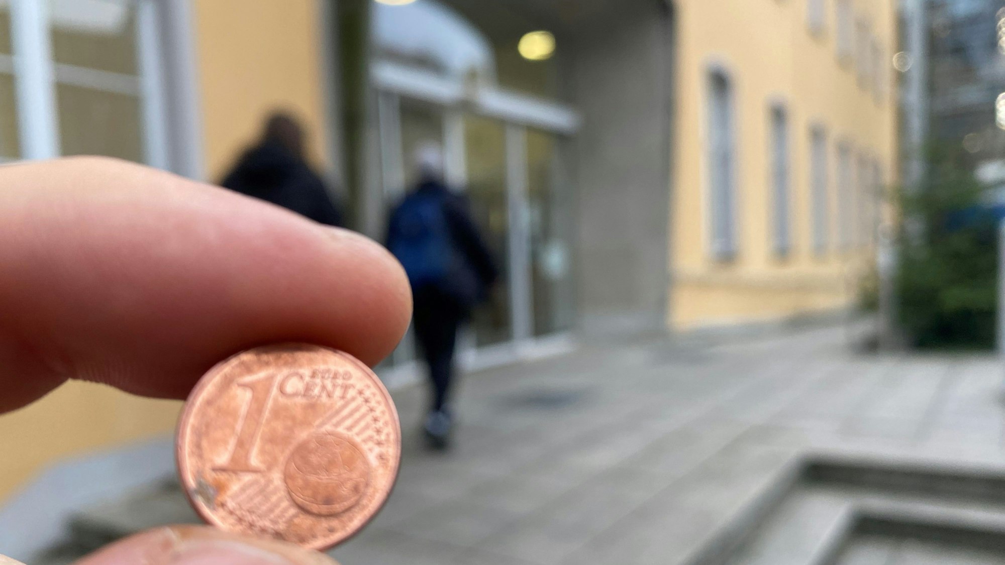 Zu sehen ist eine Ein-Cent-Münze und im Hintergrund das Brühler Rathaus.