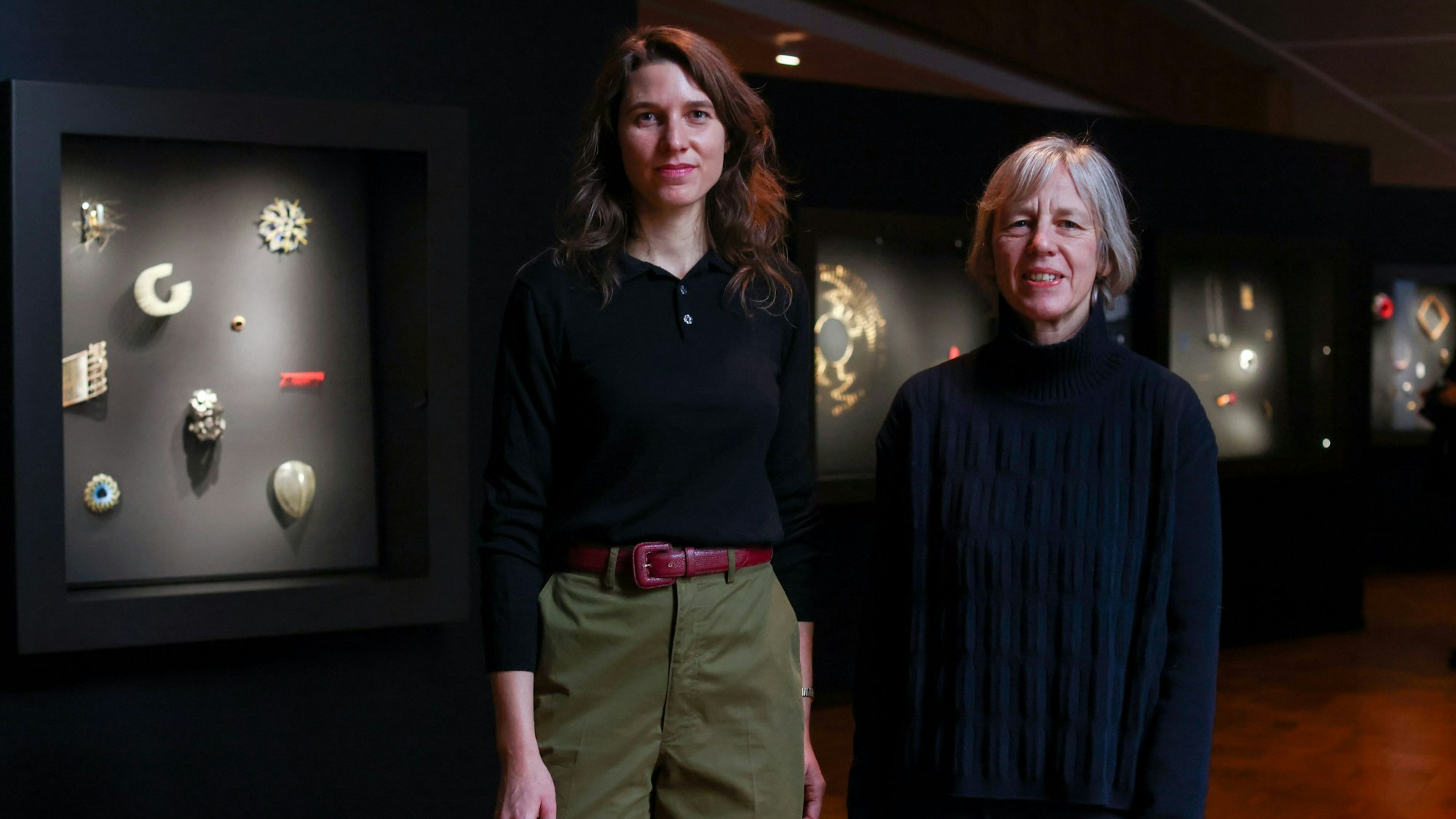 Lena Hoppe und Petra Hesse stehen vor den erleuchteten Vitrinen der Schmuckausstellung im Museum für Angewandte Kunst Köln.