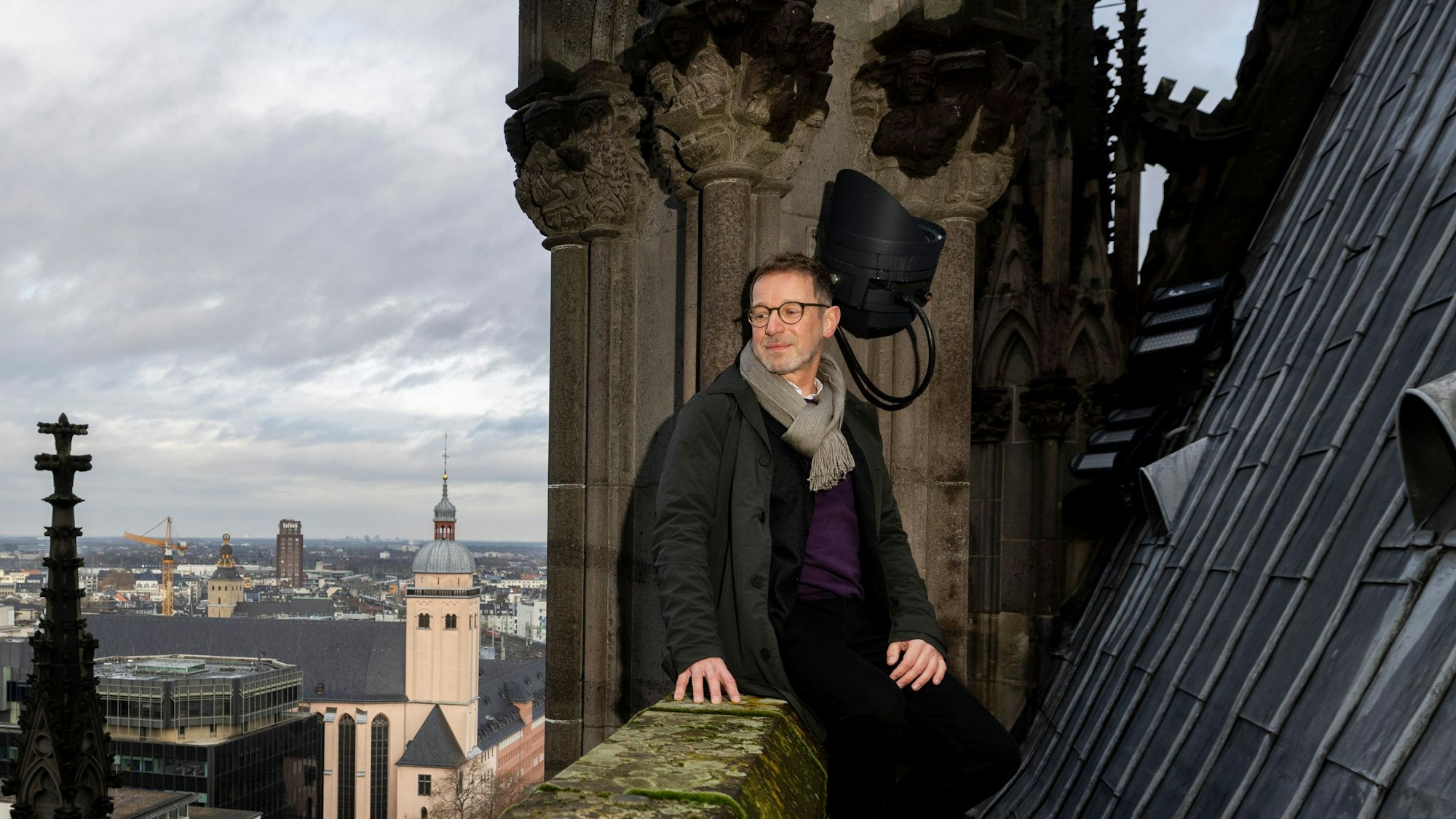 Dombaumeister Peter Füssenich sitzt auf der Brüstung des Dachumgangs am Nordquerhaus des Kölner Doms. Am Pfeiler hinter ihm Porträtdarstellungen von Mitarbeitern der Dombauhütte