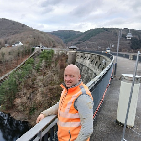 Gerrit Jaspers trägt eine orangefarbene Warnweste und steht auf der Dammkrone der Urftstaumauer.