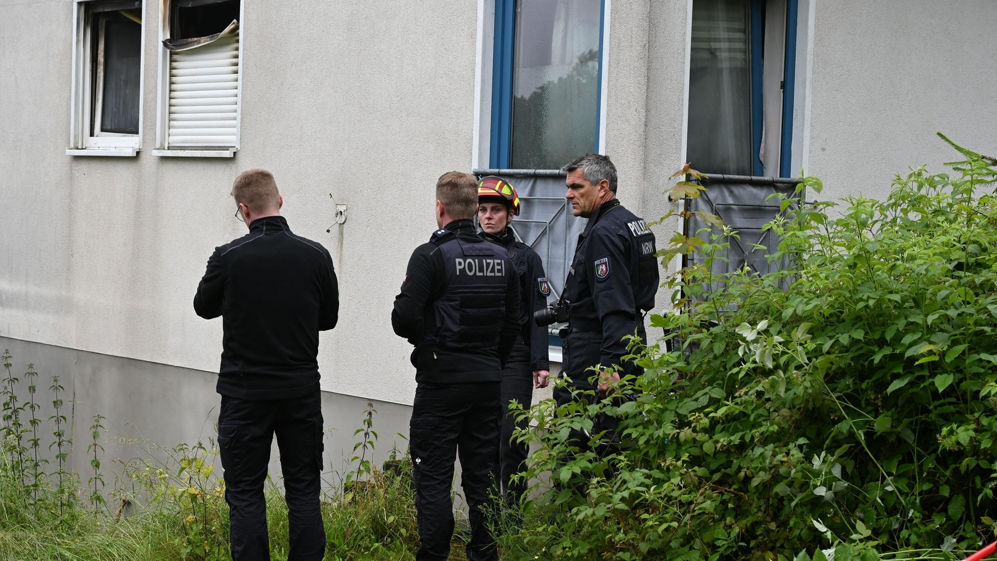 Drei Männer und eine Frau stehen vor einer Hauswand. Ein Rollladen ist durch Flammen zerstört worden.